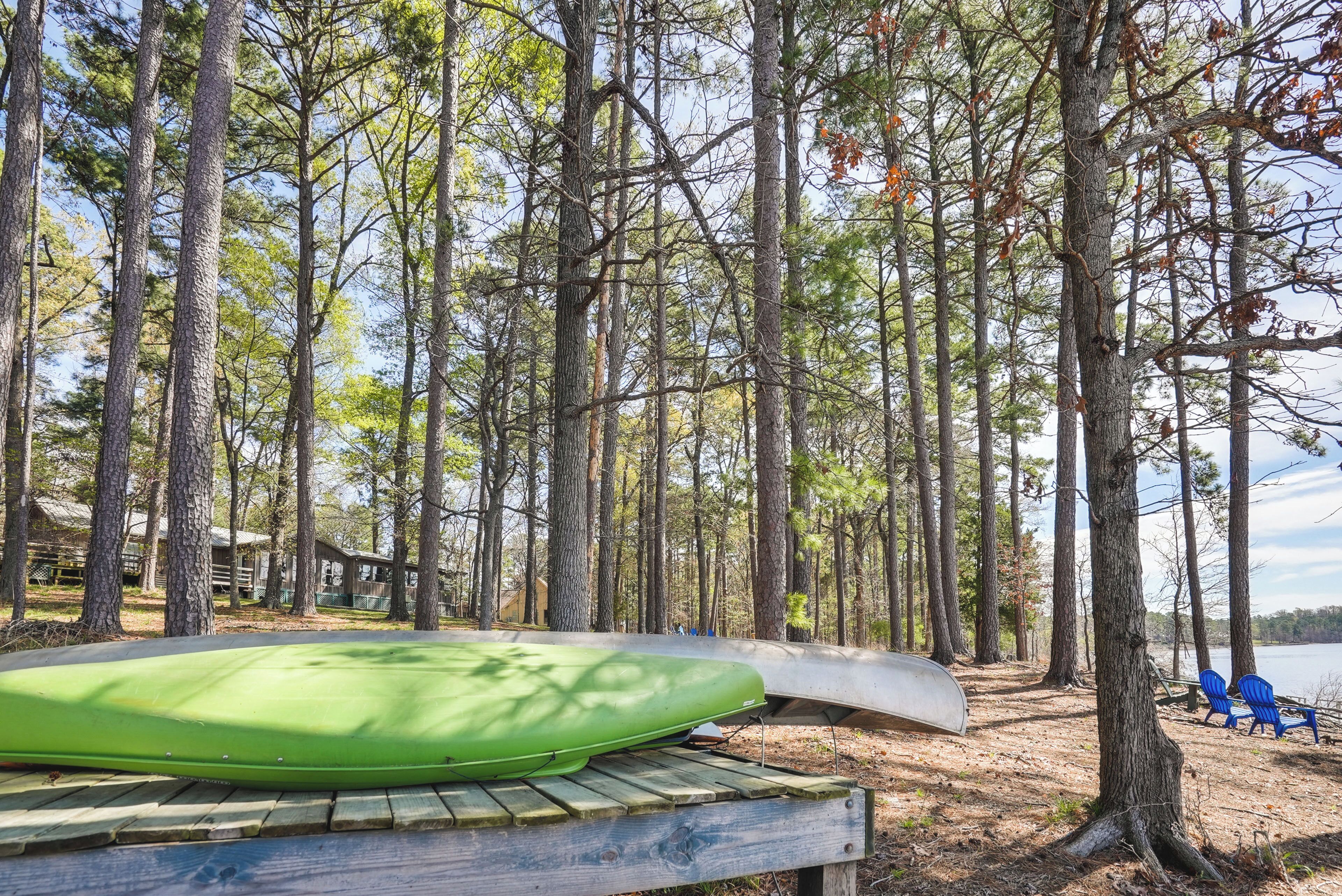 Une terrasse en bois avec un kayak vert et un canoë, entourée de grands pins et offrant une vue sur le lac.