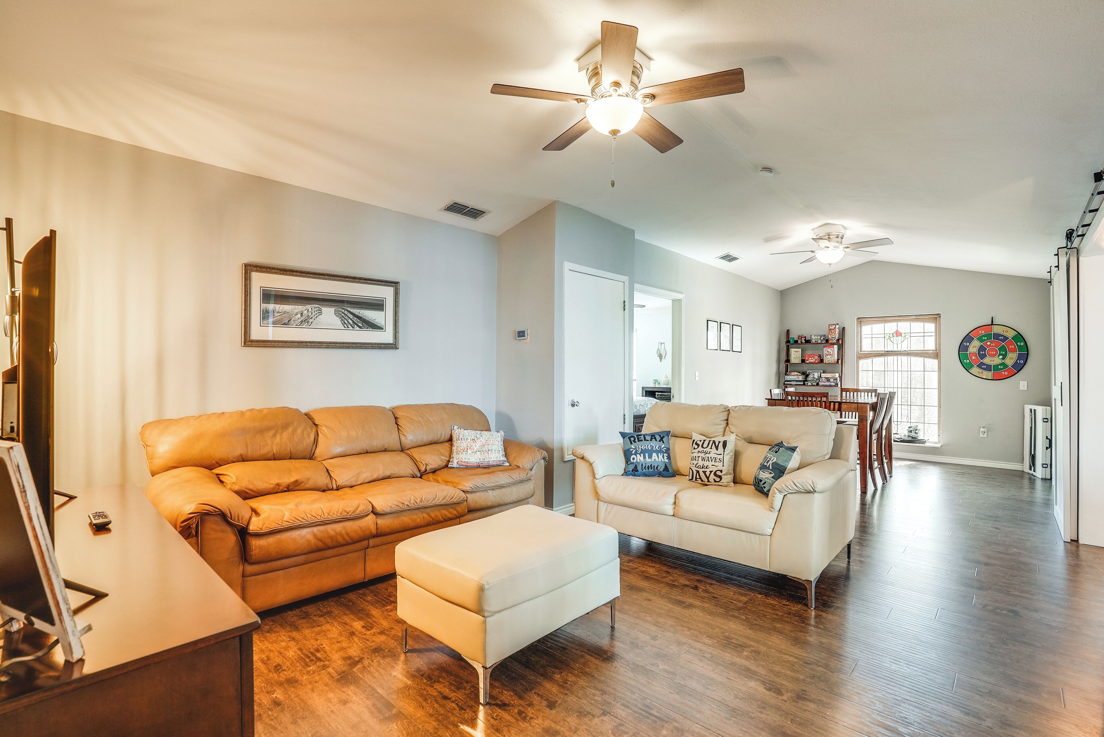A spacious living room with a brown leather sofa, a white ottoman, and a beige sofa with decorative pillows. There is a ceiling fan, a framed picture on the wall, and a dartboard.