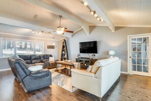 A spacious living room with a large window, a ceiling fan, a flat-screen TV, and a white sofa.