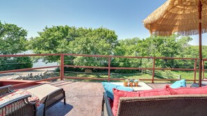 A balcony with wicker furniture, a thatched umbrella, and a view of a lake and greenery. - Scenic Great Falls Retreat On Missouri River! (Great Falls)