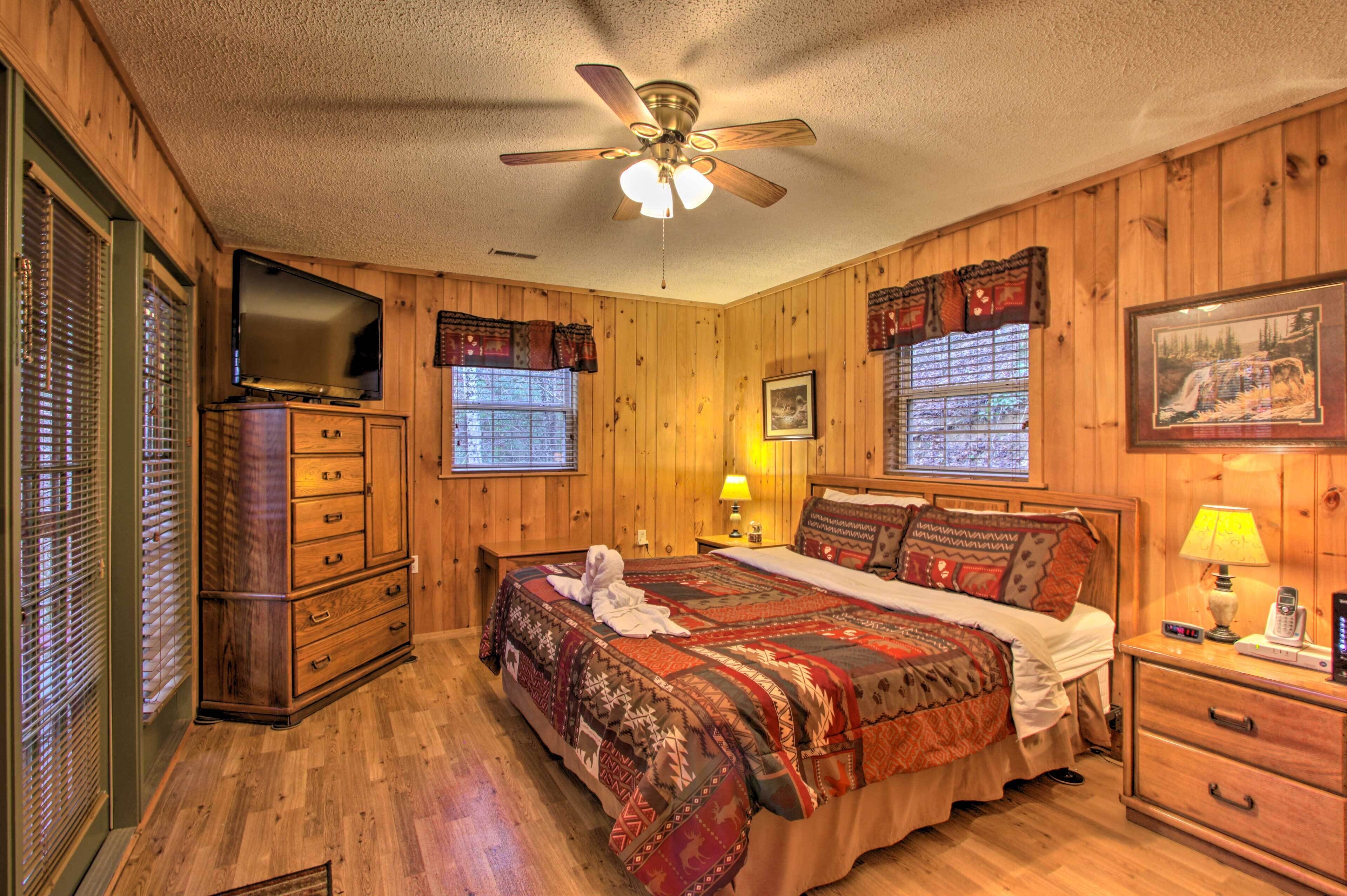 A wooden-paneled bedroom with a bed, a television, a ceiling fan, and a window with blinds.