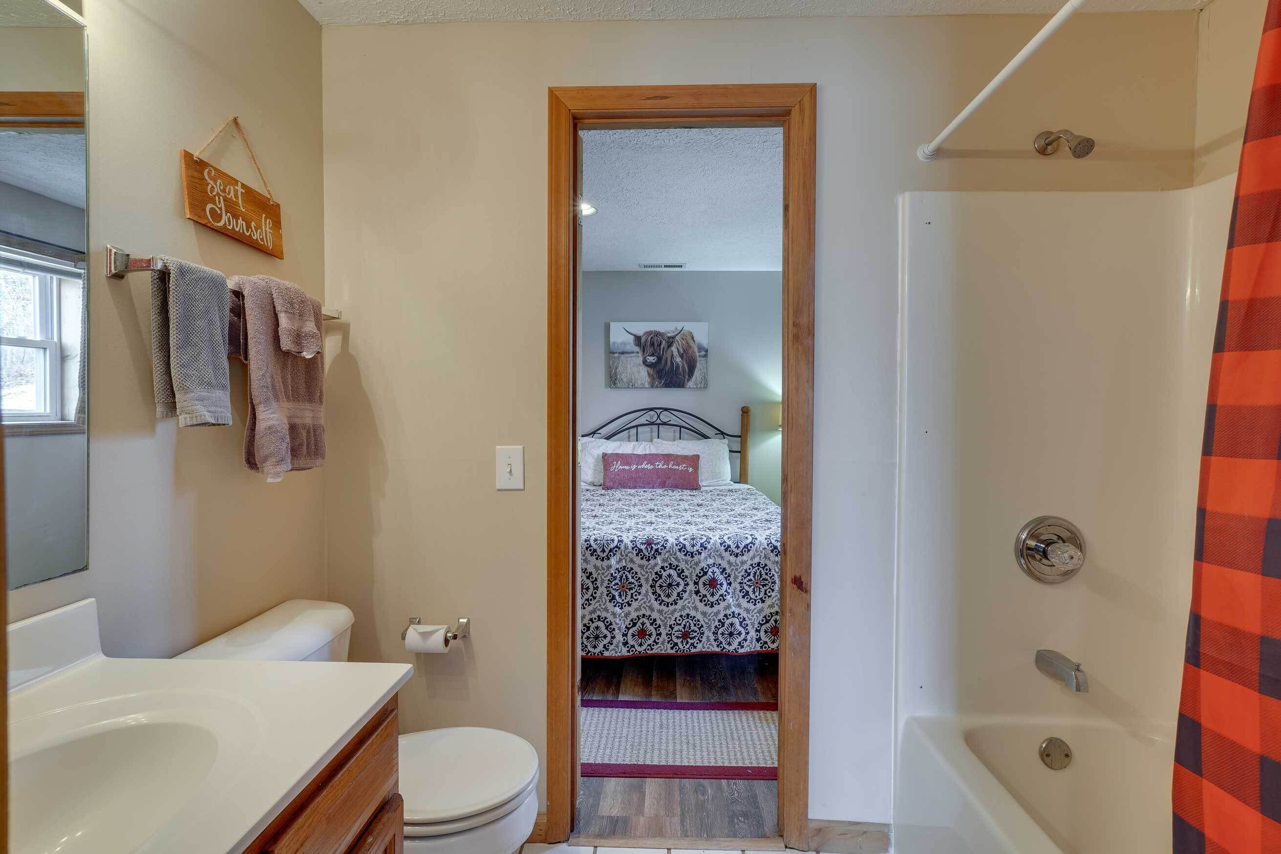 A bathroom with a shower, toilet, and sink. There is a towel rack with towels, a bed with a patterned bedspread, and a sign that says "eat yourself."
