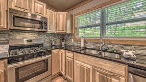 A kitchen with wooden cabinets, a stainless steel stove, a microwave, and a large window with blinds. - Ruidoso Cabin w/ Patio - Walk to Midtown! (Ruidoso)