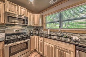 A kitchen with wooden cabinets, a stainless steel stove, a microwave, and a large window with blinds. - Ruidoso Cabin w/ Patio - Walk to Midtown! (Ruidoso)