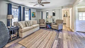 A living room with a beige sofa, patterned armchairs, a wooden floor, a ceiling fan, and a large window with curtains.