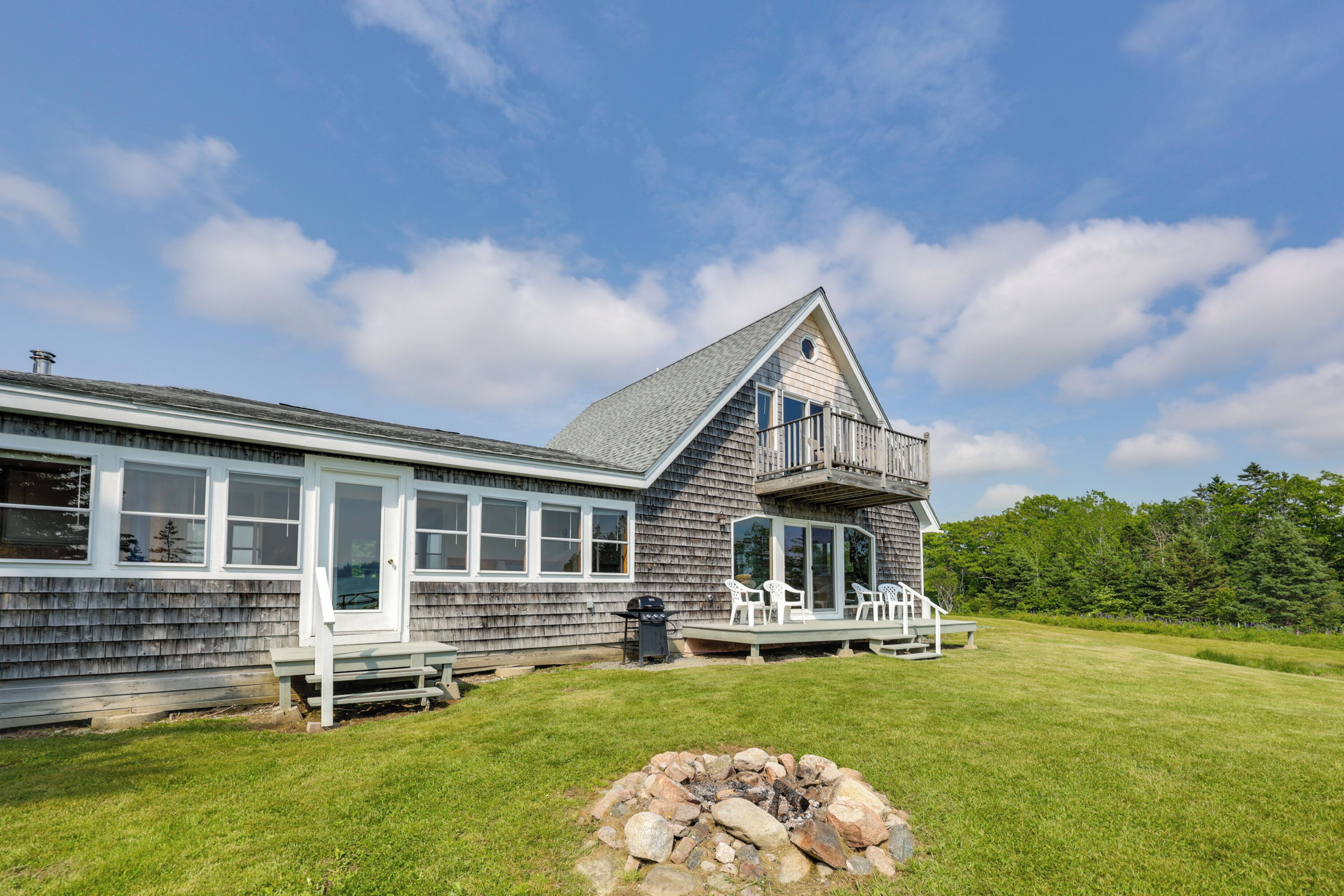 A two-story house with a large deck, a stone fire pit, and a grassy yard.