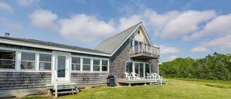 A two-story house with a large deck, a stone fire pit, and a grassy yard.