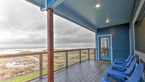 A covered deck with a view of the ocean, wooden chairs, and a door. - Sanderling Sea Cottages, Unit 5 w/ Ocean Views! (Waldport)