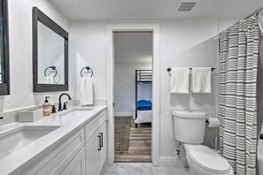 Modern bathroom with a white sink, toilet, and shower area, featuring a light-colored rug and a bed in a shared space.
