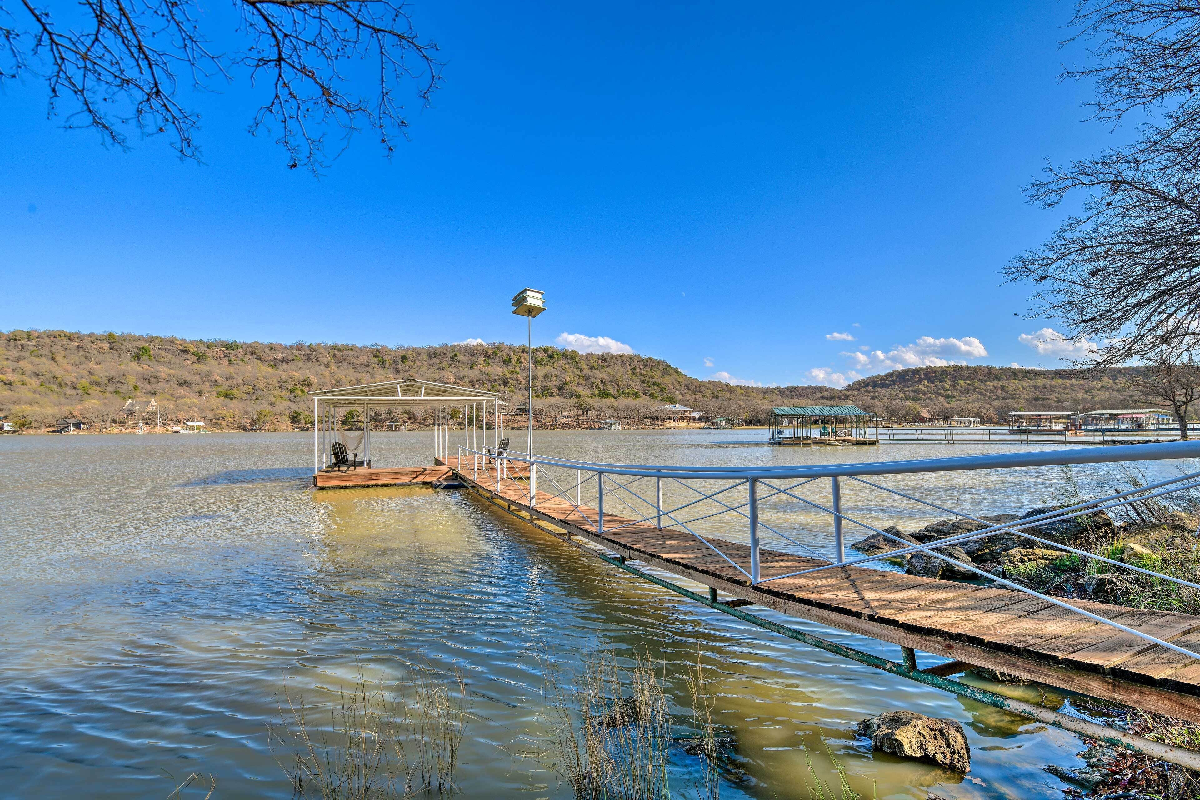 A wooden dock extending into a calm lake with a gazebo and chairs, surrounded by hills and a clear sky.