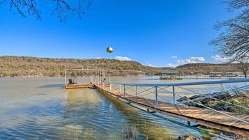 A wooden dock extending into a calm lake with a gazebo and chairs, surrounded by hills and a clear sky.