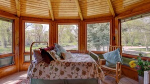 Ein gemütliches Blockhaus mit Holzinnenraum, einem Bett mit floraler Bettwäsche, einem Schaukelstuhl und großen Fenstern mit Blick auf den umliegenden Wald.