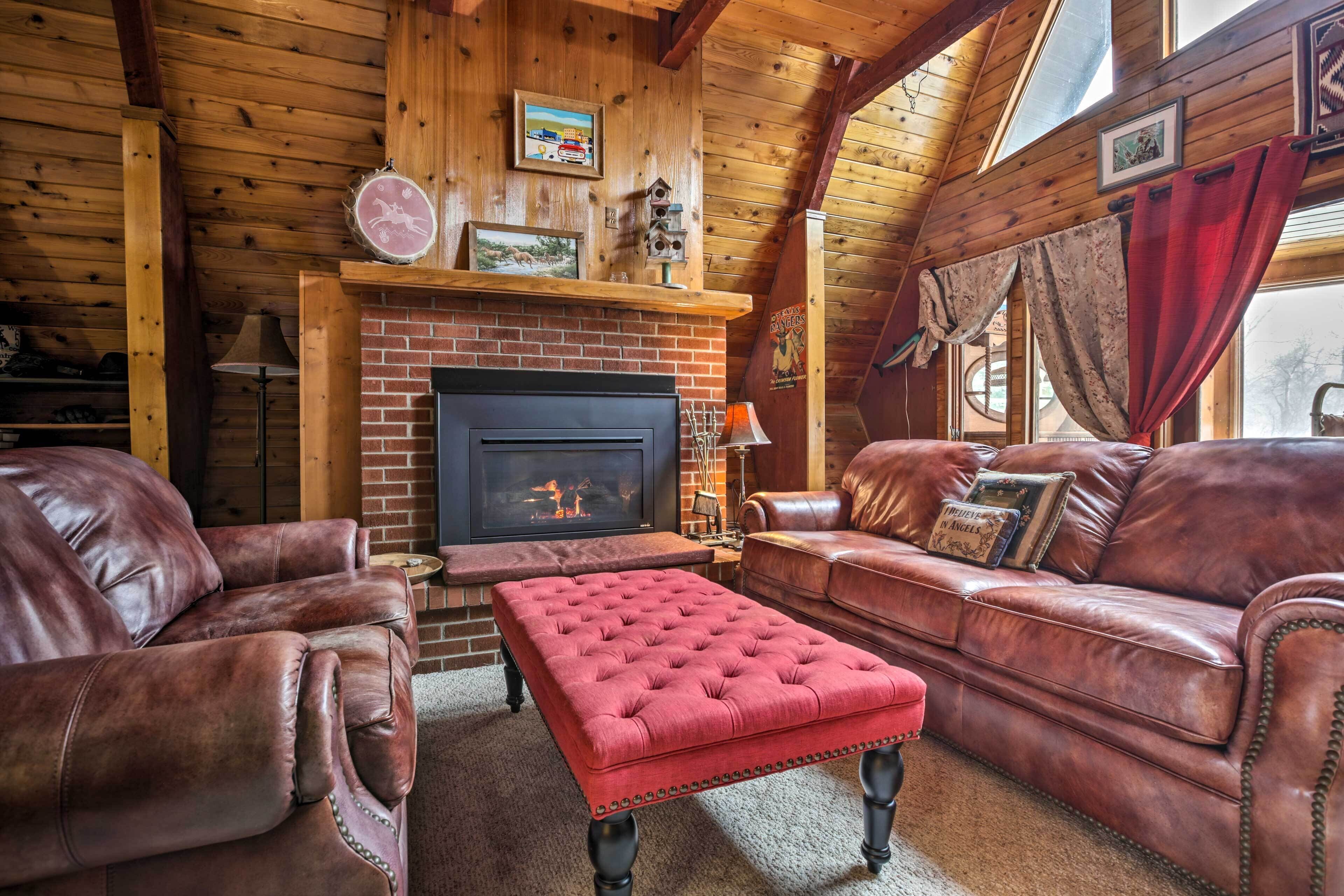 A cozy log cabin interior with a fireplace, leather sofas, and a red ottoman.