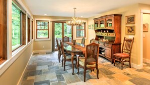 A spacious dining area with a large table and chairs, a wooden cabinet with glass doors, and a view of the outdoors through large windows.