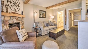 A cozy living room with a stone fireplace, a large wall clock, and a comfortable seating area with a sofa and ottoman. - Updated Onekama Resort Condo on Portage Lake! (Onekama)