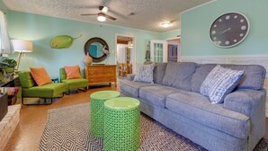 Un salon avec un canapé bleu, des poufs verts, une horloge ronde et un ventilateur de plafond.
