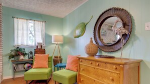 Une pièce avec un fauteuil vert, une commode en bois, un miroir rond et une fenêtre avec des rideaux.