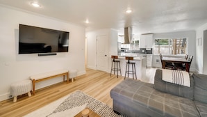 Una sala moderna con un sofá gris, un banco de madera y un televisor montado en la pared. Se ve una cocina con comedor al fondo.