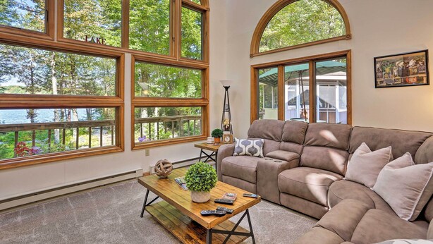 A living room with large arched windows, a brown leather sofa, a wooden coffee table, and a view of trees and a lake. - Pocono Lake Paradise w/ Private Beach & Dock! (Pocono Lake)