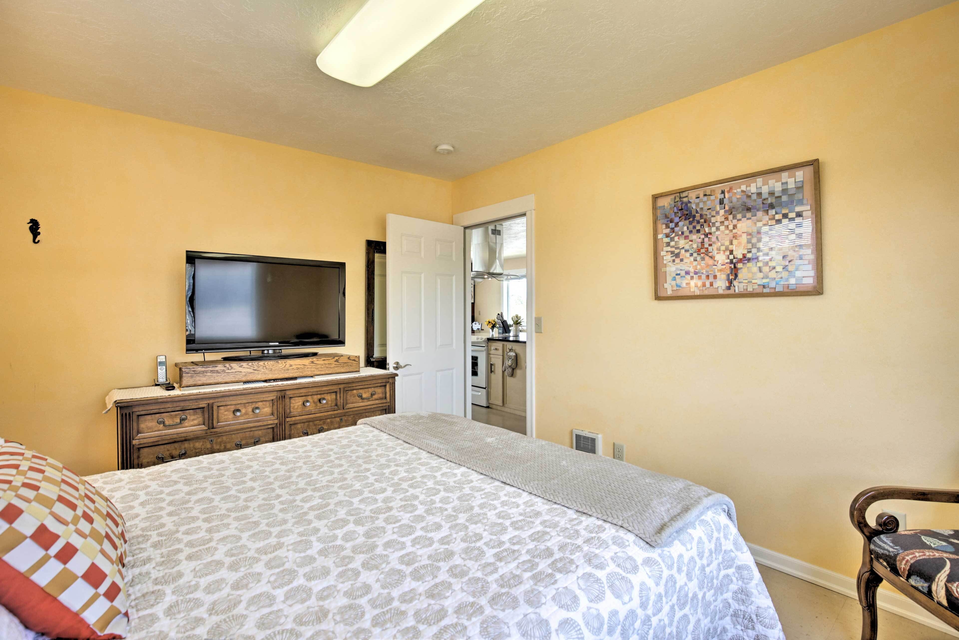 Un dormitorio con una cama, un televisor sobre un aparador, una puerta que da a una cocina y un cuadro en la pared.