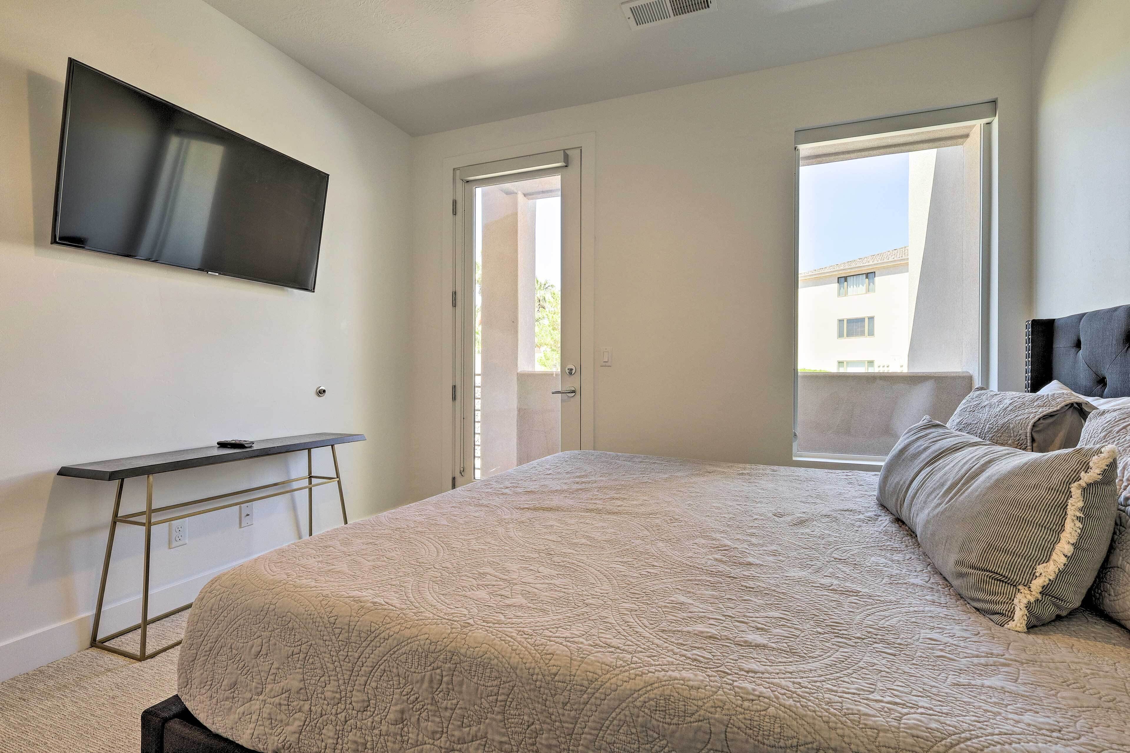 A bedroom with a bed, a TV mounted on the wall, a desk, and a window.