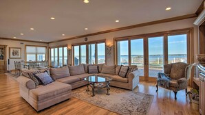 A spacious living room with a large sectional sofa, a glass coffee table, and a view of the ocean through large windows. - Waterfront Home w/ Large Deck + Private Pool! (Westhampton Beach)