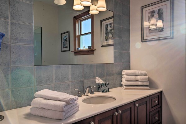 A bathroom with a large mirror, a sink, and a window. There are towels stacked on the counter and a tissue box. - Waterfront Home w/ Large Deck + Private Pool! (Westhampton Beach)