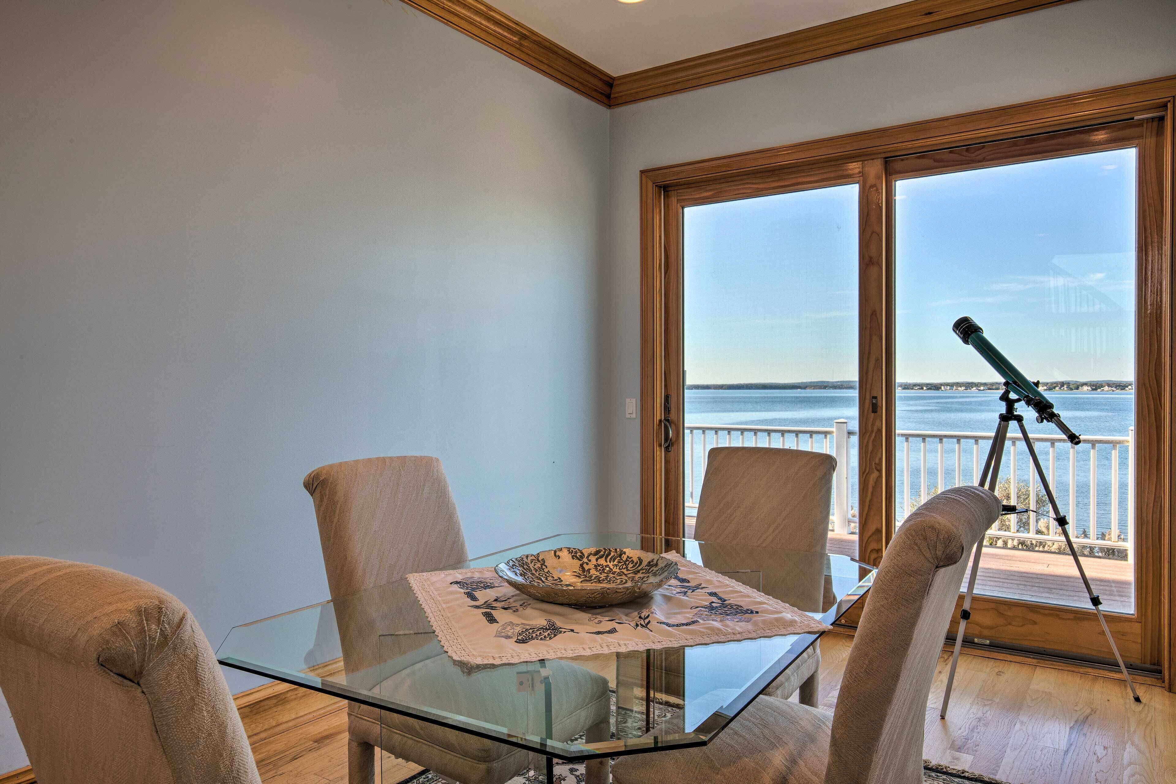 A glass-top table with a decorative bowl, two beige chairs, and a telescope on a balcony with a view of the sea.
