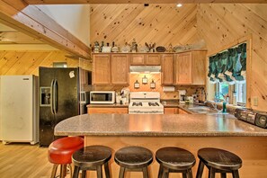 Une cuisine avec des meubles de rangement en bois, un plan de travail en granit, un réfrigérateur, un four à micro-ondes, une cuisinière et trois tabourets.