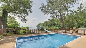 Une piscine rectangulaire avec un toboggan bleu et une échelle, entourée d’une terrasse en bois et d’arbres.