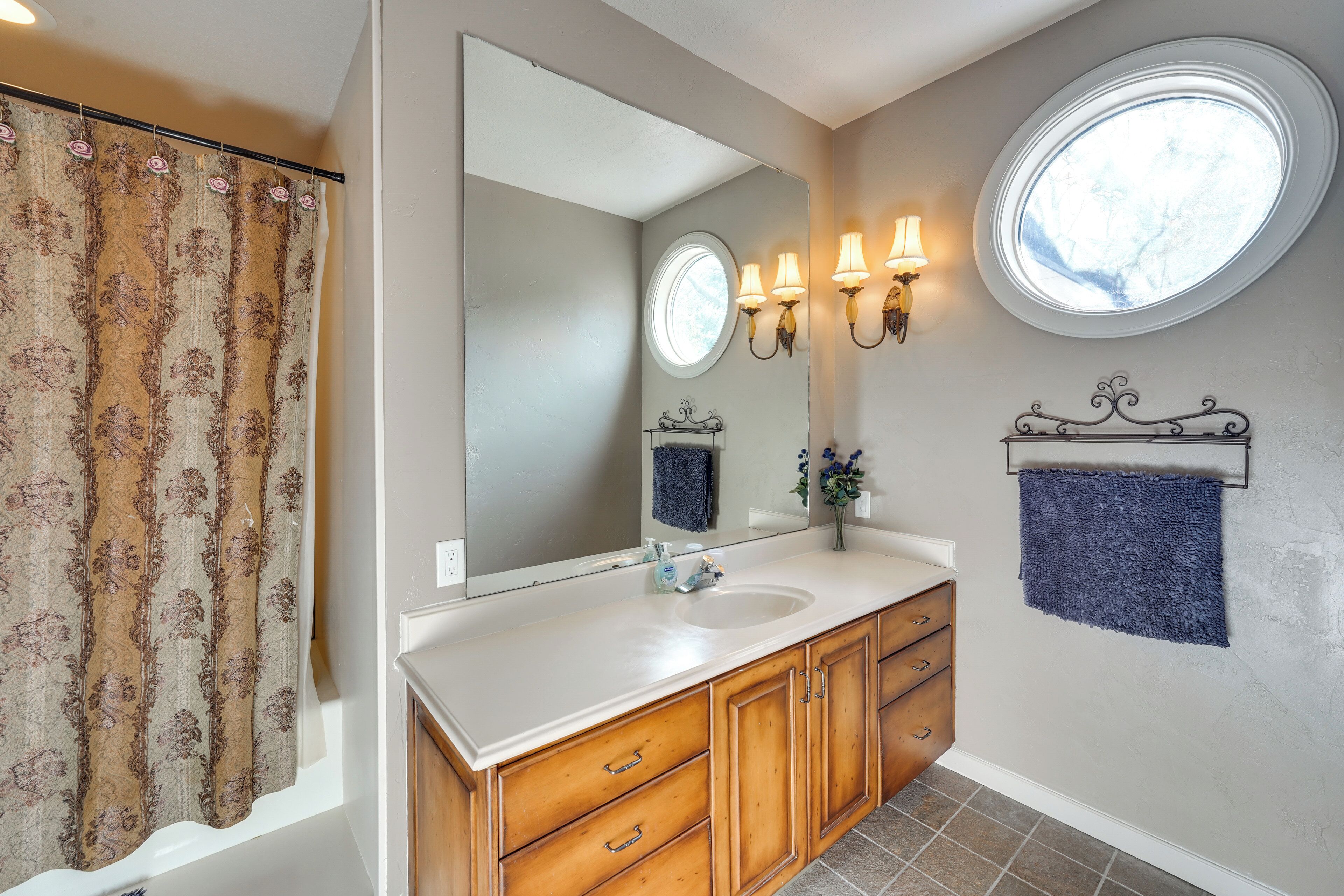 A bathroom with a large mirror, two wall-mounted lights, a window, and a towel rack.