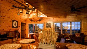 A cozy cabin interior with wooden walls, a dining table with woven placemats, a fireplace, and a large window offering a view of the outdoors.
