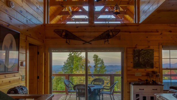 A wooden cabin interior with a large tree branch chandelier, a dining area with chairs, and a view through the glass doors.