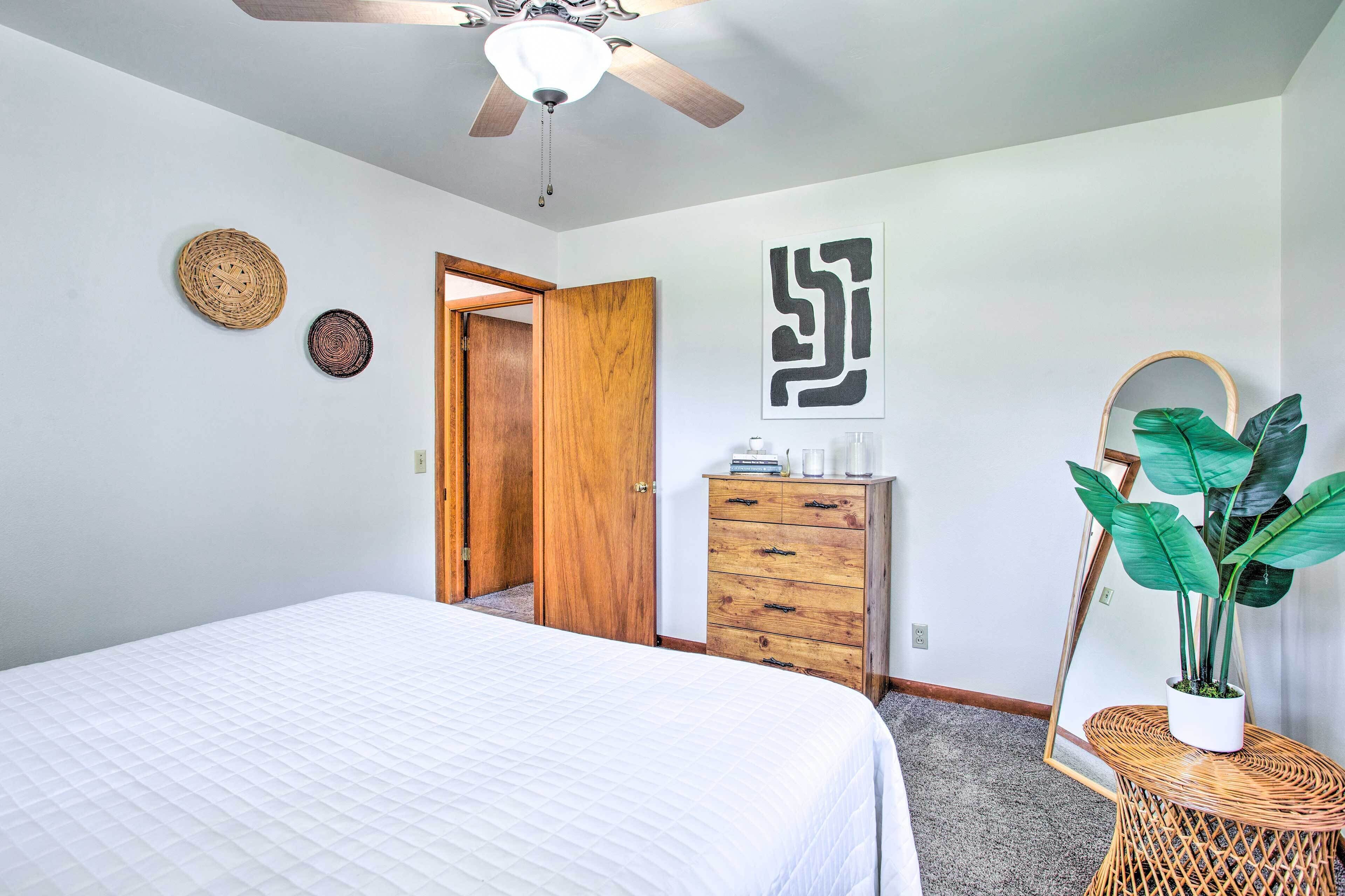 A bedroom with a bed, a ceiling fan, a wooden wardrobe, a dresser, a mirror, and a potted plant.