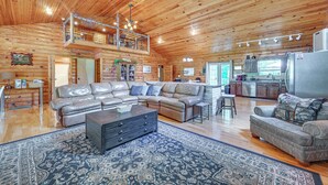 A spacious living area with a large sectional sofa, a dark wooden coffee table, and a patterned rug. A kitchen area is visible in the background. - Kentucky Group Getaway w/ Dock + Lake Access (Burnside)