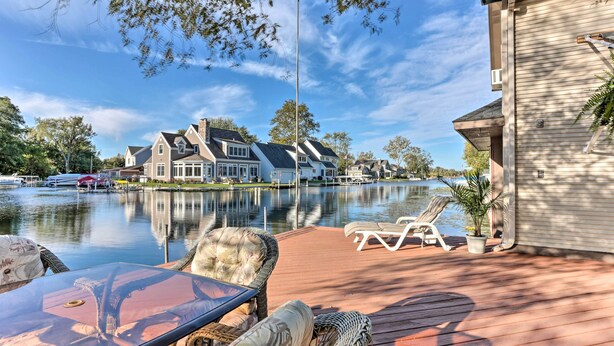 A lakeside deck with wicker furniture, a glass table, and a view of houses across the water. - Waterfront Syracuse Home: Deck, Fire Pit & Kayaks (Syracuse)
