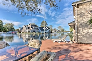 A lakeside deck with wicker furniture, a glass table, and a view of houses across the water. - Waterfront Syracuse Home: Deck, Fire Pit & Kayaks (Syracuse)