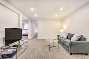 A modern living room with a flat-screen TV on a glass table, a grey sofa with patterned cushions, and a small round coffee table. - Bright & Modern Arvada Home w/ Yard & Patio (Westminster)