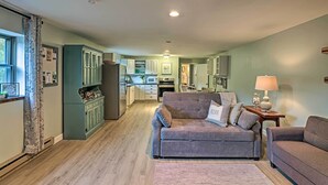 A spacious living room with a grey sofa, a smaller armchair, and a wooden coffee table. A kitchen with white cabinets and a microwave is visible in the background. - Newly Renovated Penn Yan Apartment < 1 Mi to Lake! (Penn Yan)