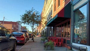 A street view with parked cars, a sidewalk, and outdoor seating at a cafe.