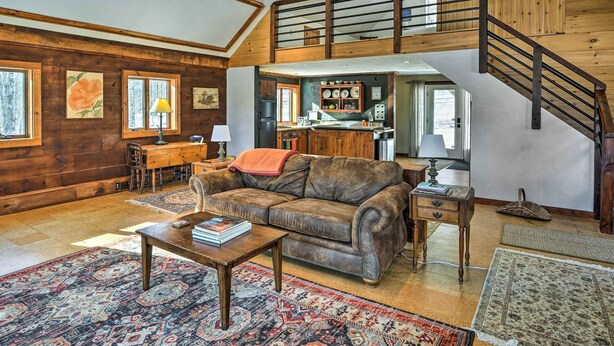 Uma sala de estar ampla com um sofá de couro, mesa de centro de madeira e uma área de jantar com mesa e cadeiras de madeira. Uma cozinha com armários de madeira e uma escada que leva a um andar superior também são visíveis.