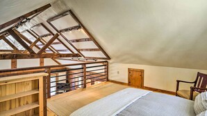 Quarto com teto inclinado, vigas de madeira, cama, cadeira e uma pequena janela.