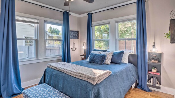 A bedroom with a large bed, blue curtains, a ceiling fan, and a bookshelf. - Central Cottage w/ Grill, 1 Mi to Cajun Field (Lafayette)