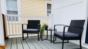 A covered porch with two black wicker chairs and a small table, a potted plant, and a white railing. - Modern, All New 3BR Near UMASS (Lowell)