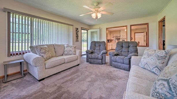 A living room with a beige sofa, two armchairs, a ceiling fan, and a view of the kitchen through an open door.