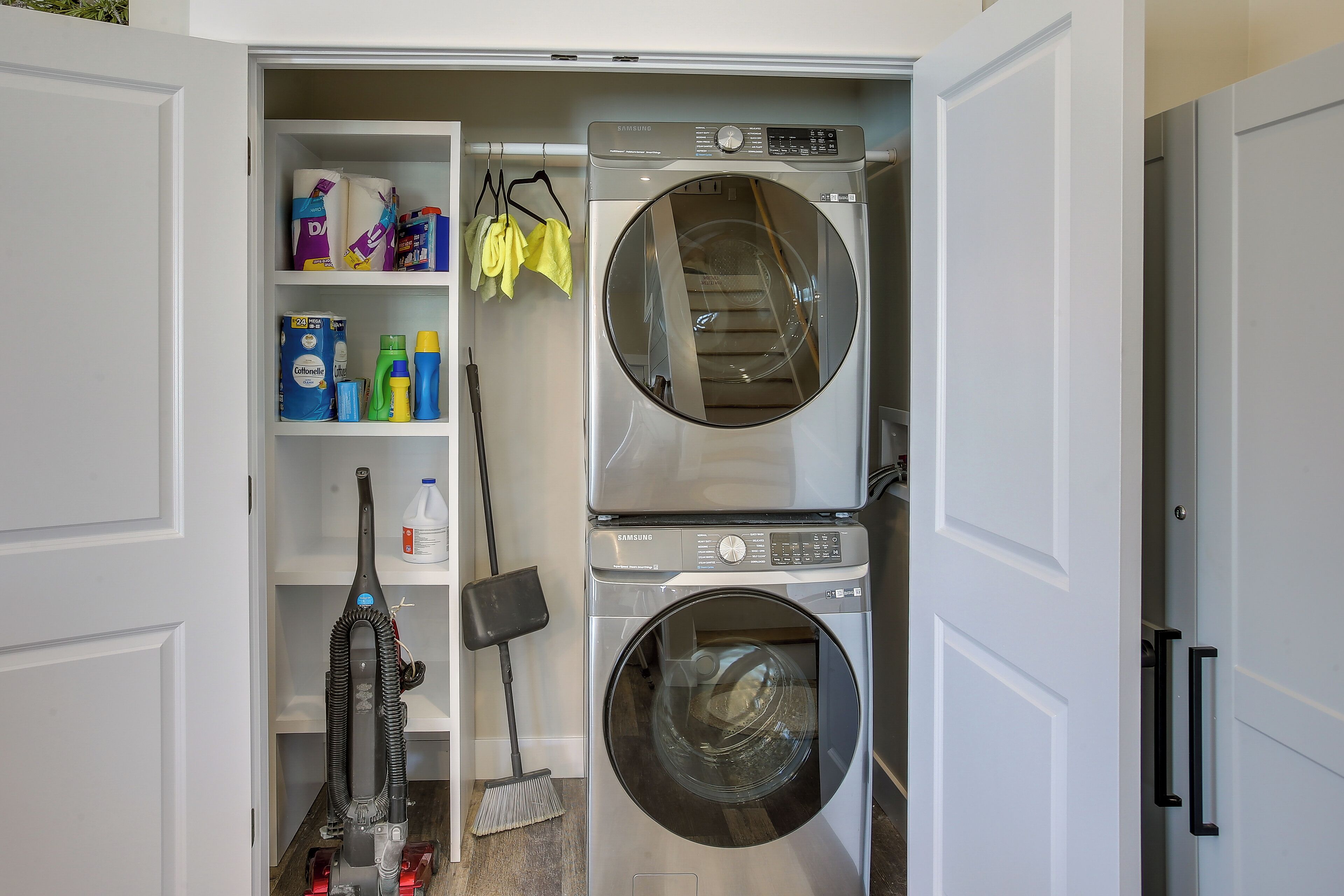 Une buanderie équipée d’un lave-linge et d’un sèche-linge à chargement frontal, d’étagères contenant des produits de nettoyage et d’un aspirateur.