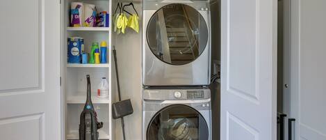 Une buanderie équipée d’un lave-linge et d’un sèche-linge à chargement frontal, d’étagères contenant des produits de nettoyage et d’un aspirateur.