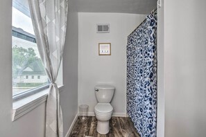 A bathroom with a toilet, a window with curtains, a shower curtain, and a light fixture on the wall. - Charming Indianapolis Home ~ 4 Mi to Dtwn! (Indianapolis)