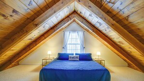 A bedroom with a sloped ceiling, a bed with a blue bedspread, two bedside tables with lamps, and a window with curtains.