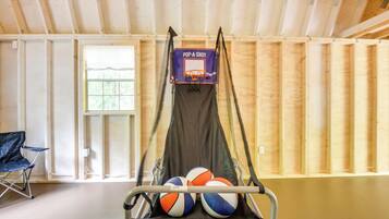 Un aro de baloncesto con tres balones en un soporte, una silla plegable azul y una ventana con vistas a árboles.